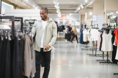 Tarz sahibi bir adam perakende mağazasında elbise raflarını karıştırıyor, modern bir alışveriş merkezinde satın alma kararları alıyor.