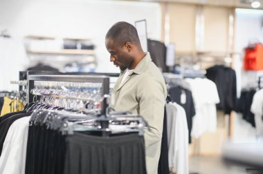 Afro-Amerikalı bir adam alışveriş merkezinde elbise raflarını karıştırıyor, şık kıyafetler seçiyor, modern moda ve perakende deneyimi sergiliyor.