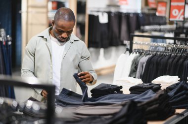 Şık bir Afro-Amerikan erkeği yeni kıyafetler alıyor, modern bir giyim mağazasında pantolonları inceliyor.