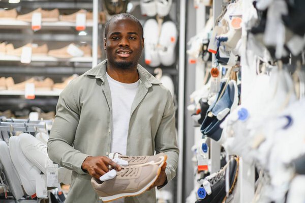 Молодой африканский американец выбирает бежевые кроссовки в обувном магазине, держит их в руках и улыбается.