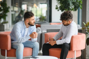 Psikolog, erkek bir hastayla terapi seansında dertlerini modern bir ofiste tartışırken not alıyor.