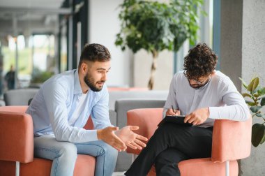 Erkek hasta, modern ofisteki terapi seansında not alan psikoloğa sorunlarını anlatıyor.