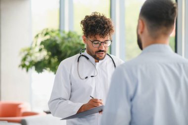 Orta Doğulu doktor, modern bir hastanede semptomları açıklayan bir hastayı dinlerken not alıyor.