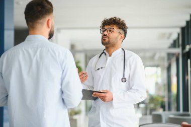 Orta Doğulu doktor elinde bir not panosuyla, hastayla tanıyı tartışıyor. Parlak, modern bir klinikte, iletişimi ve güveni arttırıyor.