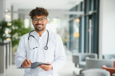 Modern bir klinikte notlar alırken gülümseyen kendinden emin bir erkek pratisyenin portresi profesyonellik ve uzmanlık gösteriyor.