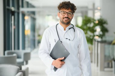 Beyaz önlüklü ve steteskoplu kendine güvenen Arap doktor hastane koridorunda dikilirken elinde bir pano tutuyor.