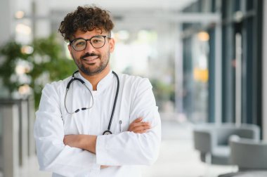 Beyaz önlük ve steteskop giyen kendinden emin bir erkek doktor. Parlak ve temiz iç mekanlarla dolu modern bir klinikte içten içe gülümsüyor.