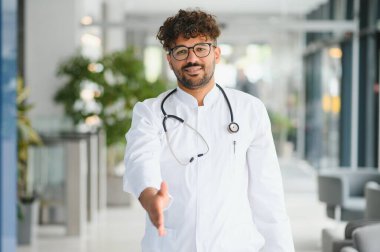 Dost canlısı Arap doktor, parlak, modern hastane koridorunda sıcak bir tokalaşma sunuyor, profesyonellik ve bakım yayıyor.