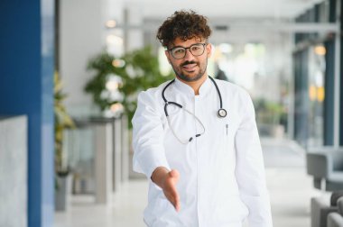 Genç bir erkek doktor el sıkışmak için elini uzatıyor. Bir hastayı sıcak bir şekilde modern bir kliniğe kabul ediyor.