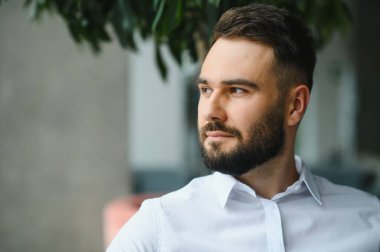 Beyaz gömlekli sakallı işadamı, modern ofis ortamında kameradan uzağa bakıyor, düşüncelere dalmış.