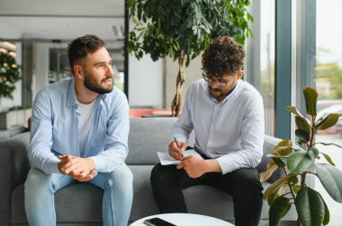 Psikolog, modern ofiste terapi seansı sırasında erkek hastaların sorunları hakkında konuşmalarını dinlerken not alıyor.