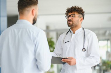 Arap doktor elinde bir dosya tutuyor ve modern, parlak bir klinik ortamında erkek bir hastaya teşhis koyuyor.