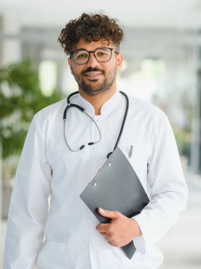 Kendine güvenen Arap doktor hastane koridorunda duruyor, elinde sağlık raporu tutuyor ve steteskop takıyor, sağlık sektöründe profesyonelliği somutlaştırıyor.