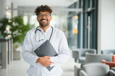 Parlak ve modern bir hastane lobisinde elinde bir pano tutarken gülümseyen kendine güvenen bir doktorun portresi profesyonellik yayıyor.