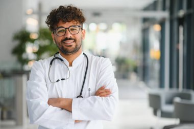 Kendine güvenen Arap doktor kollarını kavuşturup gülümsüyor laboratuvar önlüğü ve steteskop giyiyor, modern hastane koridorunda.