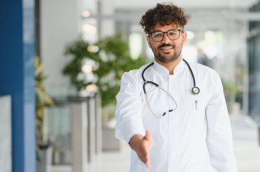 Gülümseyen Arap doktor el sıkışmak için elini uzatıyor, modern klinikte hastaları ağırlıyor, sağlık ve güven sunuyor.