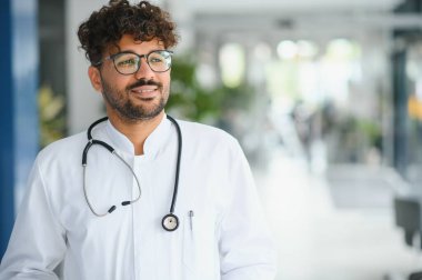 Hastane koridorunda gülümseyen kendine güvenen Arap doktor, modern tıp ortamında sağlık, profesyonellik ve uzmanlığı temsil ediyor.