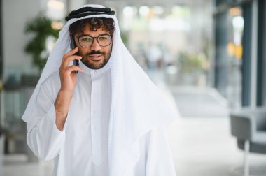 Kendine güvenen Arap işadamı geleneksel kandura giyiyor modern ofis binasında akıllı telefon kullanıyor.
