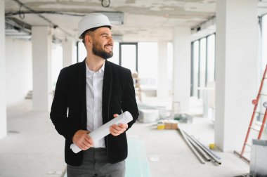 Kendine güvenen mimar kasketli ve elinde planlar tutarak modern binanın inşaat alanındaki ilerlemeyi kontrol ediyor.