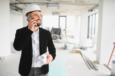 Telefonla konuşan ve inşaat alanının planlarını elinde tutan kasketli gülümseyen bir mimar.