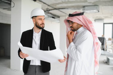 İnşaat halindeki bir binanın içinde düşünceli bir Arap yatırımcının planlarını gösteren bir mimar.