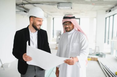 Mimar başlıklı ve Arap yatırımcı inşaat halindeki binanın planlarını inceliyor, gülümsüyor ve tartışıyorlar.