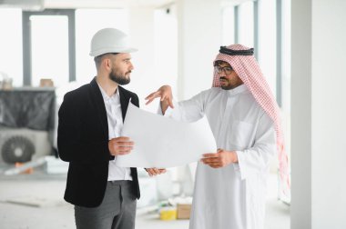 Güvenlik kaskı takan mimar, inşaat halindeki bir binanın içindeki Arap yatırımcının planlarını gösteriyor.