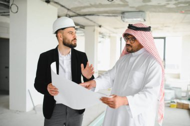 Güvenlik kaskı takan mimar, inşaat halindeki bir binanın içindeki Arap yatırımcının planını gösteriyor.