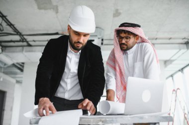 Mimar sabit şapka takıyor, Arap yatırımcı planları değerlendiriyor ve inşaat sırasında dizüstü bilgisayar kullanıyor.