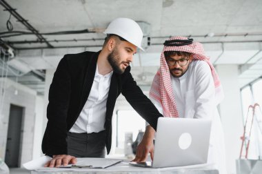 Başlık takan mimar ve yatırımcı dizüstü bilgisayardaki bina planlarını analiz ediyor, gayrimenkul inşaat projesini tartışıyorlar.