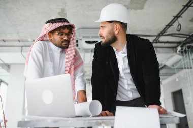 Geleneksel giysiler giyen Arap yatırımcı ve mimar inşaat halindeki bir binanın dizüstü bilgisayar ve planlarını kullanarak bir inşaat projesini tartışıyorlar.