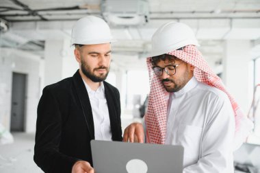 Projenin detaylarını tartışırken hareketli bir inşaat sahasında dizüstü bilgisayar kullanan miğferler.