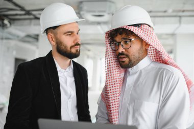 Şapkalı iki mimar inşaat alanının planlarını tartışmak için dizüstü bilgisayar kullanıyor.