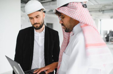 Şapkalı iki mimar inşaat sahasında dizüstü bilgisayar kullanıyor, bina planlarını tartışıyor ve proje ilerlemesini tartışıyorlar.