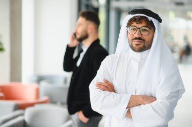 Geleneksel kandura giyen kendine güvenen Suudi işadamı modern bir ofiste müdürün telefonda konuşurken kollarını kavuşturuyor.