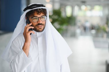 Geleneksel kandura giyen ve modern bir binada akıllı telefon kullanan Arap bir işadamının portresi.