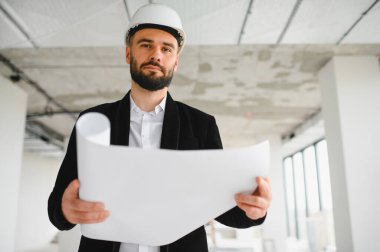 Mimar sabit bir şapka takıyor, elinde planlar tutuyor ve bir inşaat alanını inceliyor, proje geliştirme ve tasarım alanında uzmanlık gösteriyor.