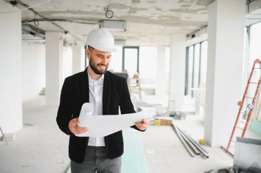 Genç mimar hareketli bir inşaat sahasının planlarını inceliyor, başlık takıyor ve kendine güvenen bir gülümseme sergiliyor.