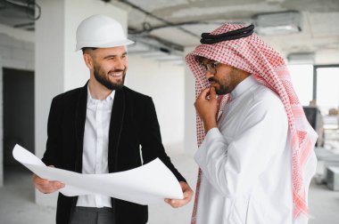 İnşaat alanındaki düşünceli Arap yatırımcıya kasketli, gülümseyen ve planlarını gösteren bir mimar.