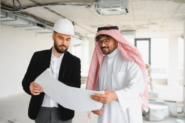 Mimar ve yatırımcı, bir inşaat sahasının içinde planları birlikte inceleyerek, yaklaşan kalkınma projesinin planlarını tartışıyor.