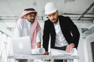 Mimar başlık takıyor ve yatırımcı geleneksel giysiler giyiyor inşaat alanının planlarını tartışıyor