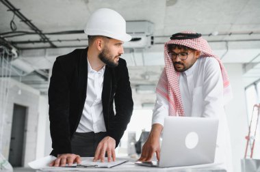 Güvenlik kaskı takan mimar ve Arap yatırımcı dizüstü bilgisayar ve planlar kullanarak gayrimenkul inşaat projesini tartışıyor