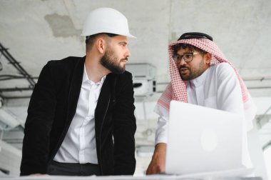 Güvenlik kaskı takan bir mimar ve Arap yatırımcı bir inşaat alanında dizüstü bilgisayarla tartışıyorlar.