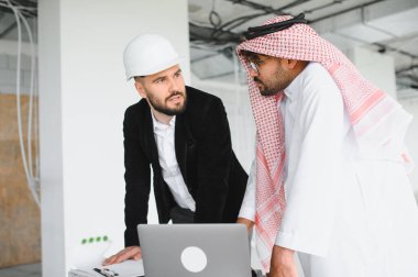 Güvenlik kaskı takan mimar ve inşaat sahasındaki bir toplantıda dizüstü bilgisayardaki bina planlarını değerlendiren Arap yatırımcı.