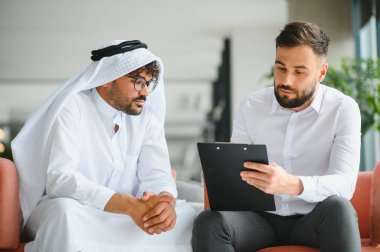 Uluslararası yönetici, Suudi iş ortağına geleneksel kandura giyen bir iş ortağıyla iş görüşmesi yaparken sözleşme detaylarını gösteriyor