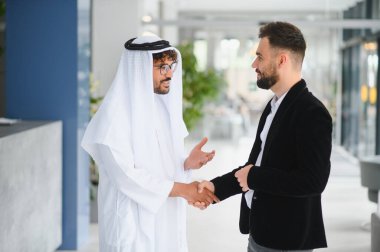 Uluslararası operasyon yöneticisi Suudi bir iş ortağıyla görüşüyor. İkisi de geleneksel kandura giyip işbirliğini teşvik ediyor.