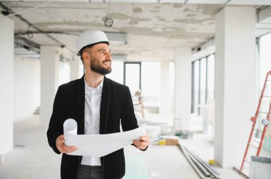 Genç mimar, yeni bir binanın inşaat alanını incelerken gülümsüyor ve elinde planlar tutuyor.