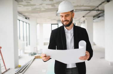 Mimar, inşaat halindeyken koruyucu miğfer takıp bina planlarını analiz ediyor.