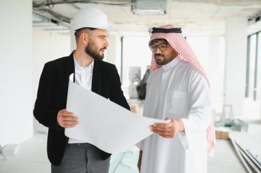 Güvenlik kaskı takan mimar, inşaat halindeki bir binanın içindeki Arap yatırımcının planını gösteriyor.