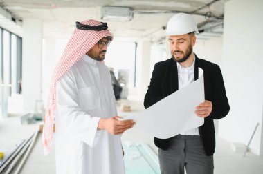 Arap yatırımcı ve bir mimar inşaat halindeki bir binanın planlarını inceliyor ve bir emlak projesinde işbirliği yaptıklarını gösteriyorlar.